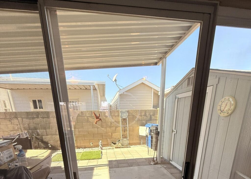 View through a glass door to a backyard patio with a shed and houses.